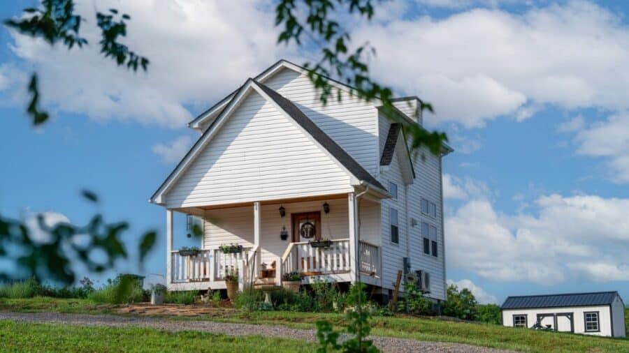 White two-story tiny farmhouse with front porch, dark gabled roof, plantings, and a matching outbuilding under blue sky