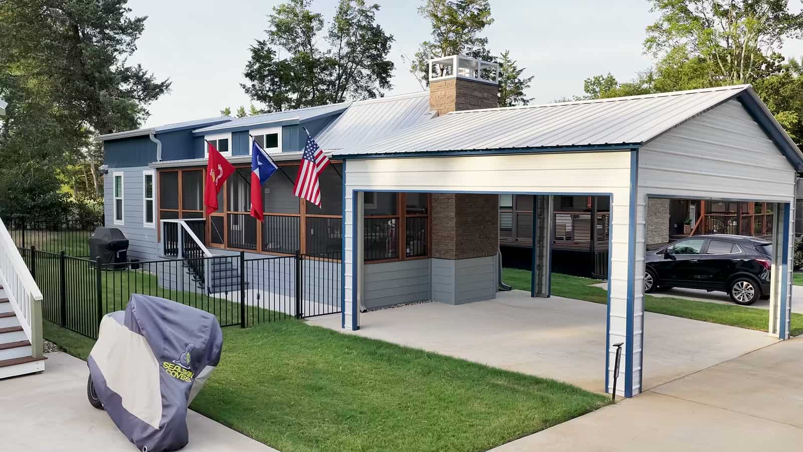Park model home with detached metal carport, fenced lawn, covered grill and SUV parked under the carport