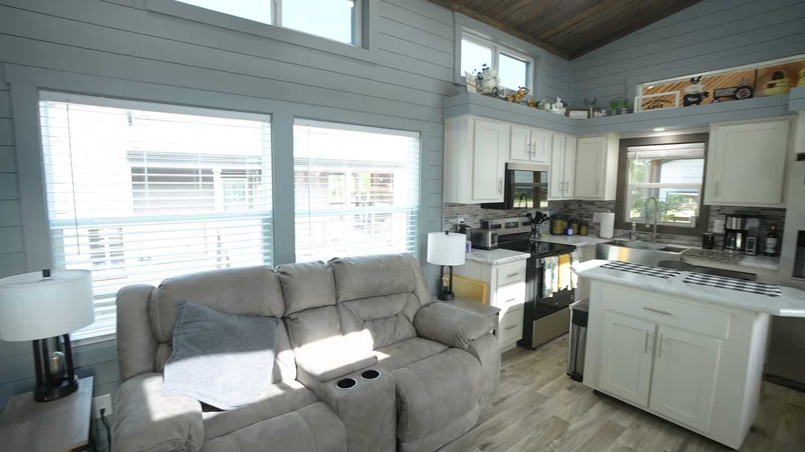 Open living room and kitchen in a park model with gray reclining sofa, large windows, island and stainless appliances