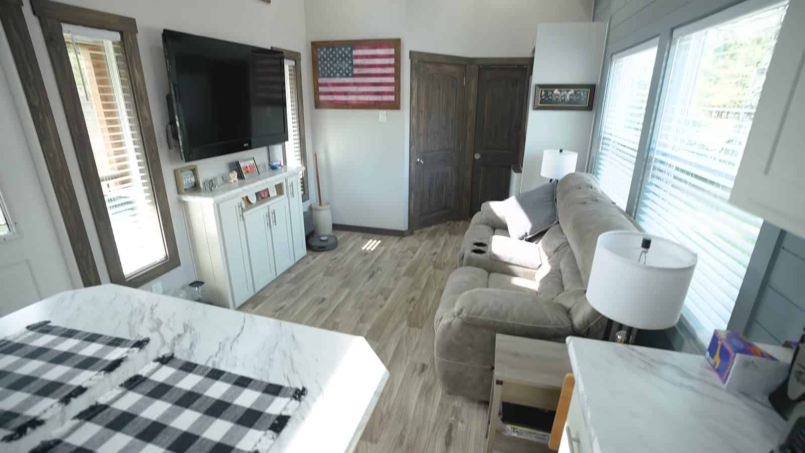 Park model living room with wall-mounted TV, framed American flag, reclining sofa, white cabinets and bedroom door