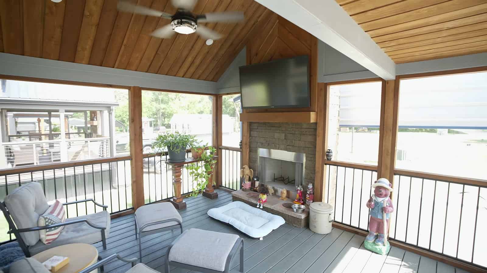 Screened porch with wood plank ceiling, ceiling fan, stone fireplace, wall-mounted TV and patio lounge chairs