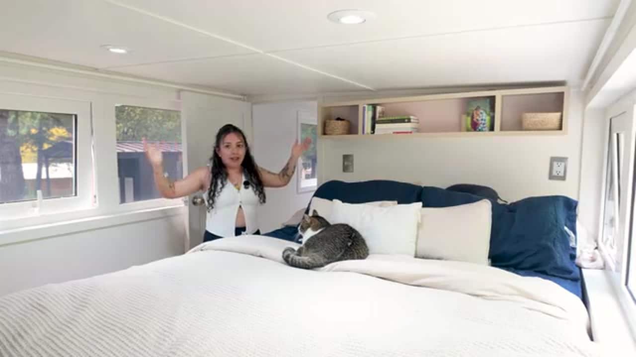 Shelby standing in the stand-up loft bedroom, arms up under the ceiling, cat on a white bed, windows on both sides