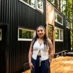Black vertical-metal tiny house on a trailer among tall trees, wood around the glass door, wood deck, Shelby standing in front