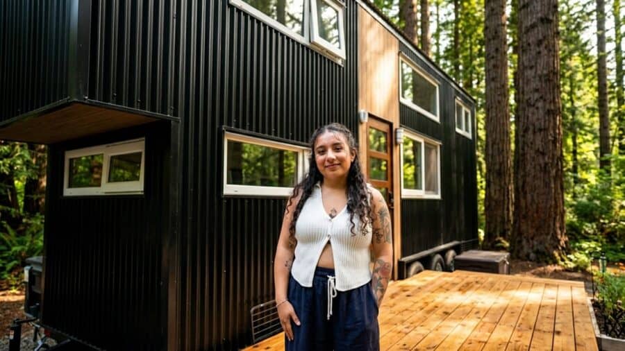 Black vertical-metal tiny house on a trailer among tall trees, wood around the glass door, wood deck, Shelby standing in front
