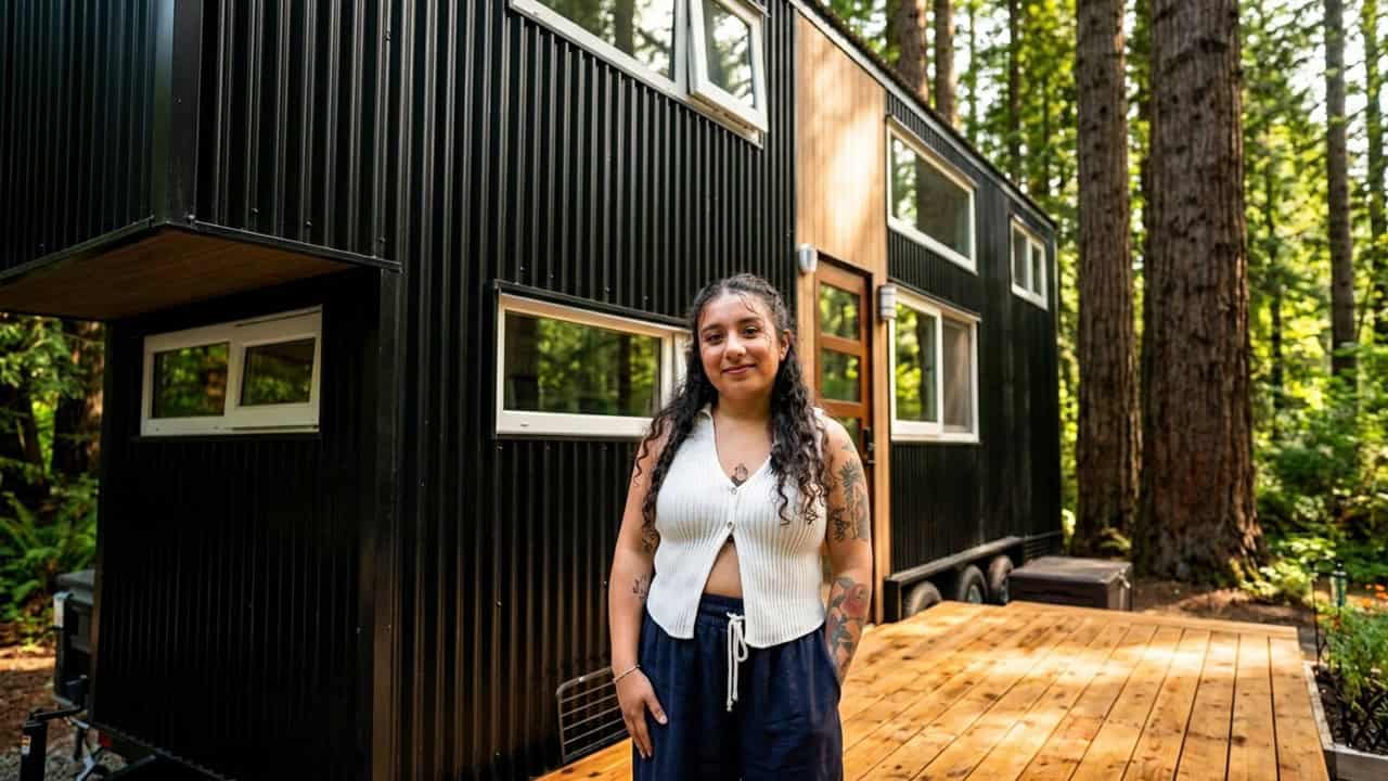 Black vertical-metal tiny house on a trailer among tall trees, wood around the glass door, wood deck, Shelby standing in front