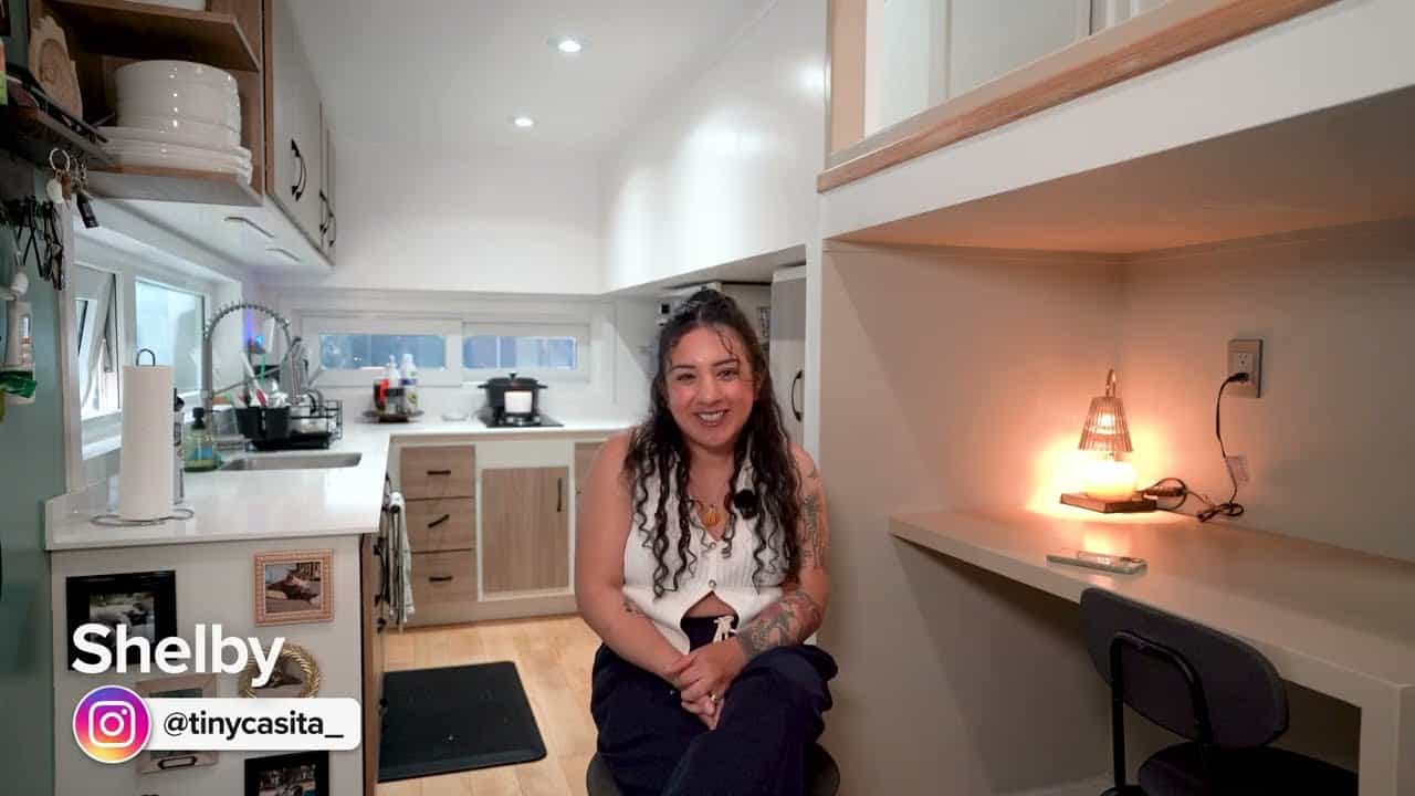 Shelby sitting in the aisle of her tiny house, kitchen to the left, under-loft desk to the right, on-screen name Shelby and Instagram @tinycasita_