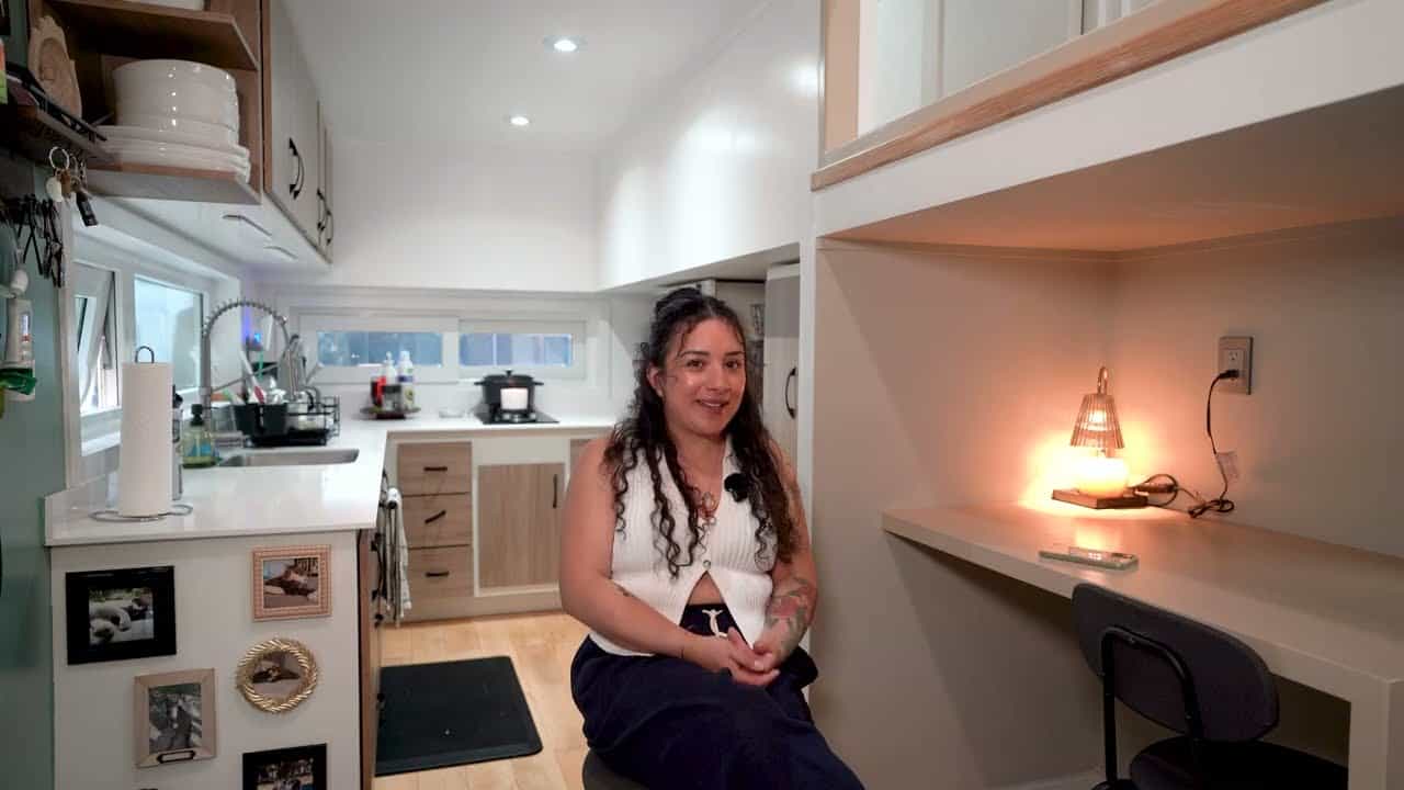 Kitchen with white counters and open wood shelves facing an under-loft desk with a lamp, Shelby seated in the aisle