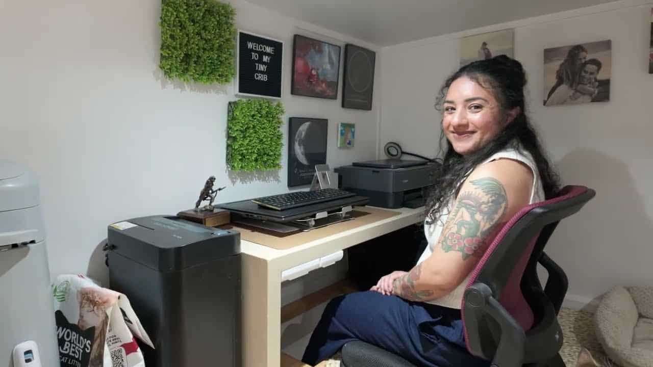 Shelby at a loft desk under a letter board that reads WELCOME TO MY TINY CRIB, moss panels and a printer on the wall