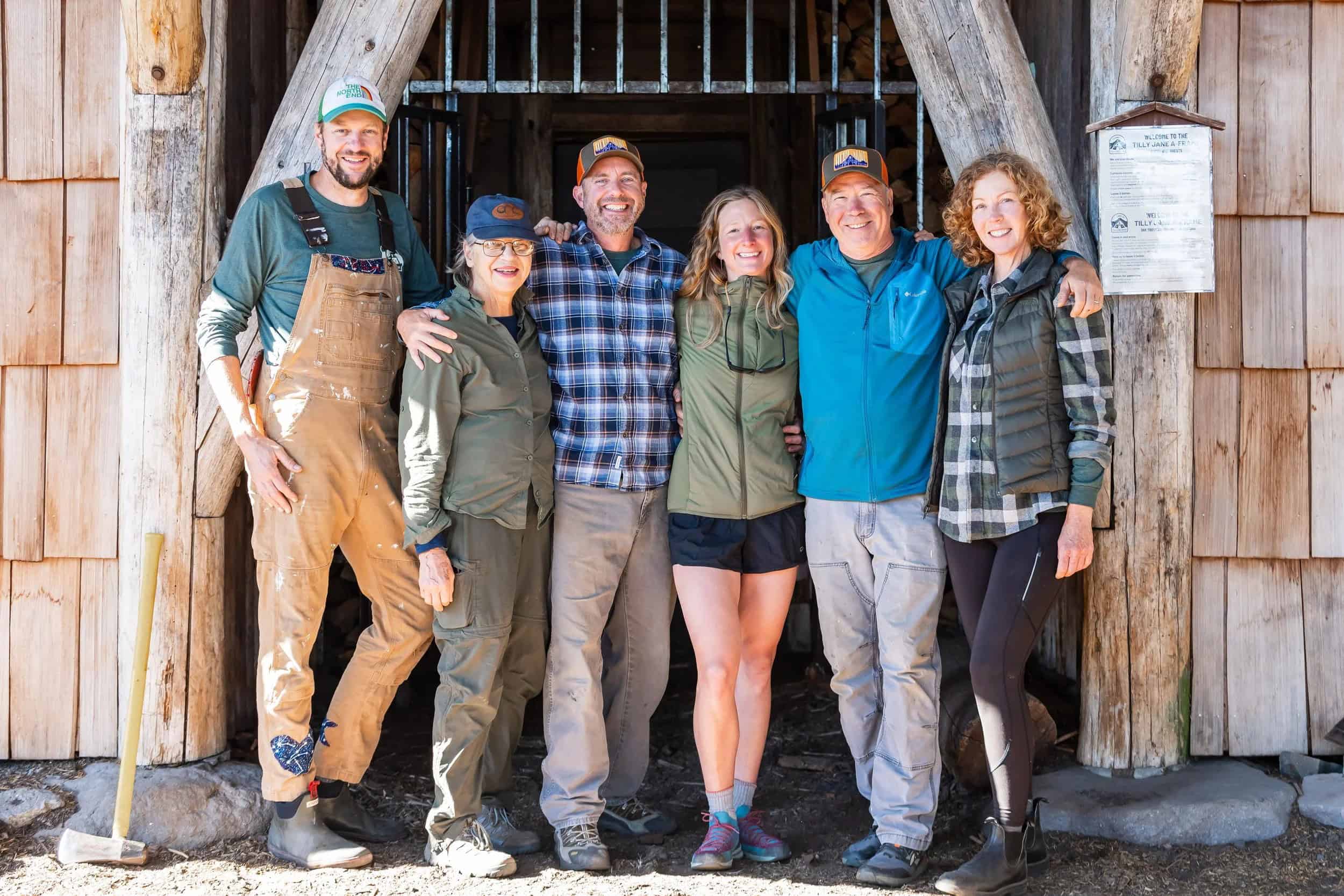 Six Friends of Tilly Jane volunteers at the A-Frame entrance, with a Welcome to the Tilly Jane A-Frame sign on a log post and stacked firewood in the alcove