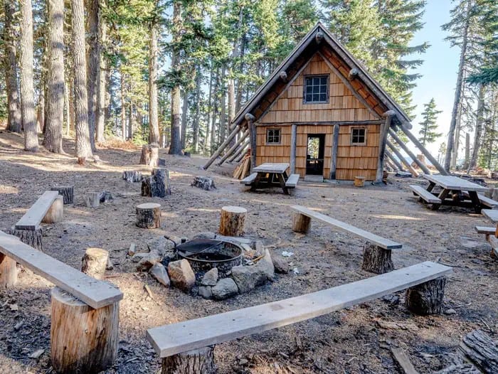 Rear summer view of the Tilly Jane A-Frame: shingled A-frame, log buttresses, a rock fire ring, stump benches, and picnic tables in the trees