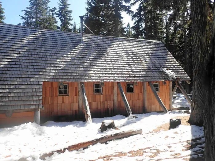 Side view of the Tilly Jane A-Frame in early winter: steep shingle roof, log posts, four small windows, a flue, and patchy snow on the needles