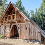 Tilly Jane A-Frame in summer: steep shingle roof, log balcony over a recessed entrance, stone foundation, and tall evergreens on Mt. Hood