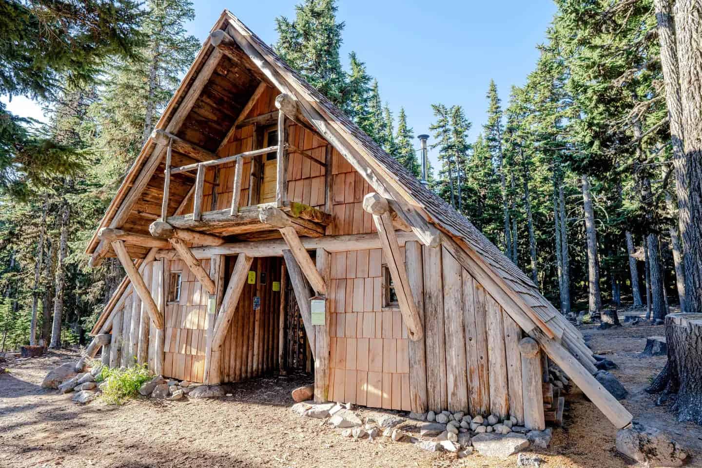 Tilly Jane A-Frame in summer: steep shingle roof, log balcony over a recessed entrance, stone foundation, and tall evergreens on Mt. Hood