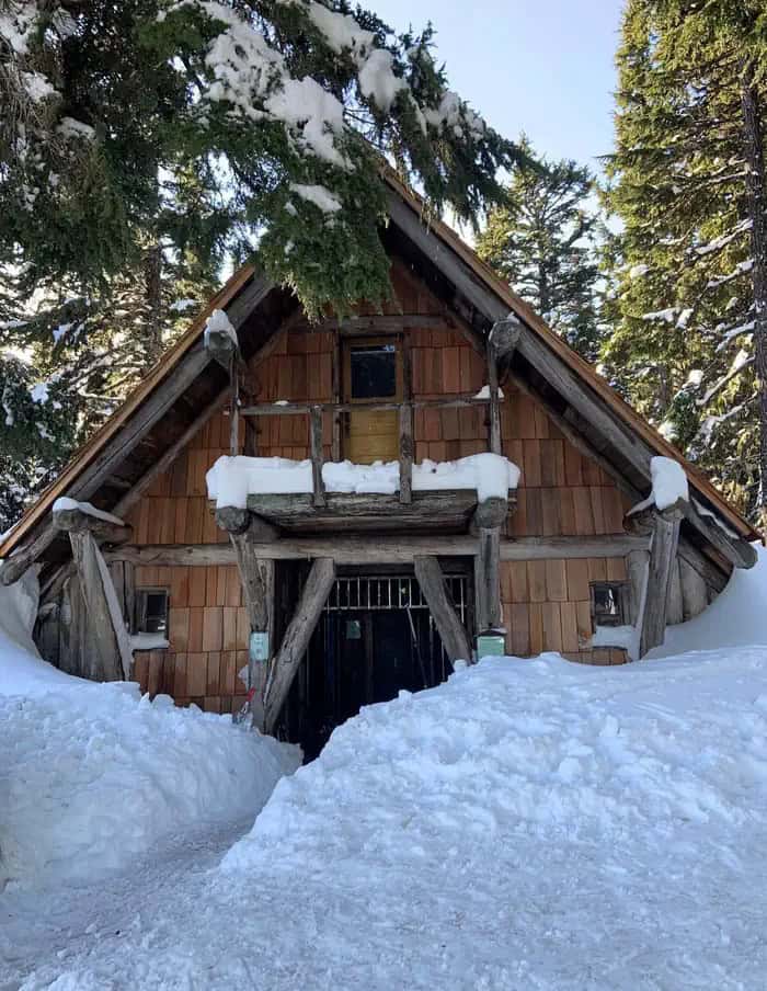 Winter view of the Tilly Jane A-Frame entrance: snow on the balcony and roof, a cleared path to the recessed door, evergreens loaded with snow