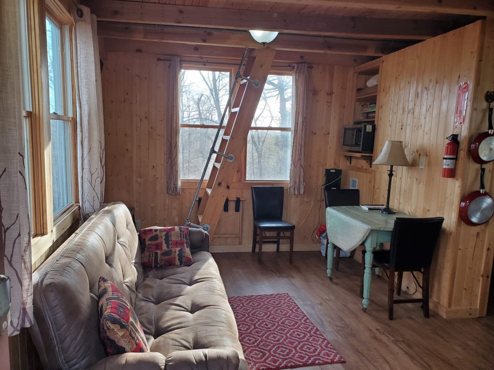 Main-floor interior with sofa, table, loft ladder, and kitchenette microwave inside the White Oak Treehouse