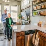 Kitchen and living in Tiny Treasure: dark-wood cabinets, light counter, grey sofa, Ben and Cheryl in the aisle