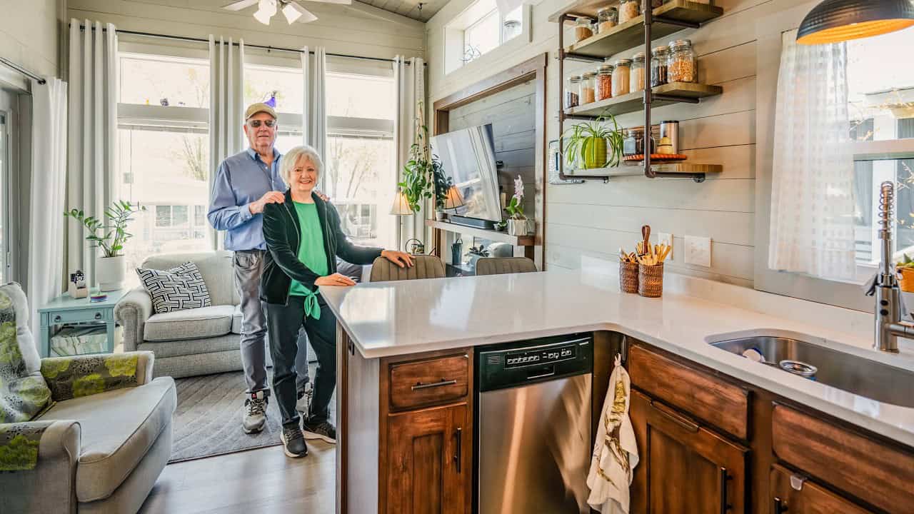 Kitchen and living in Tiny Treasure: dark-wood cabinets, light counter, grey sofa, Ben and Cheryl in the aisle
