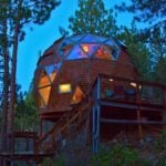 Cedar-shingle geodesic dome on a hillside timber deck at dusk, warm light in the triangular windows