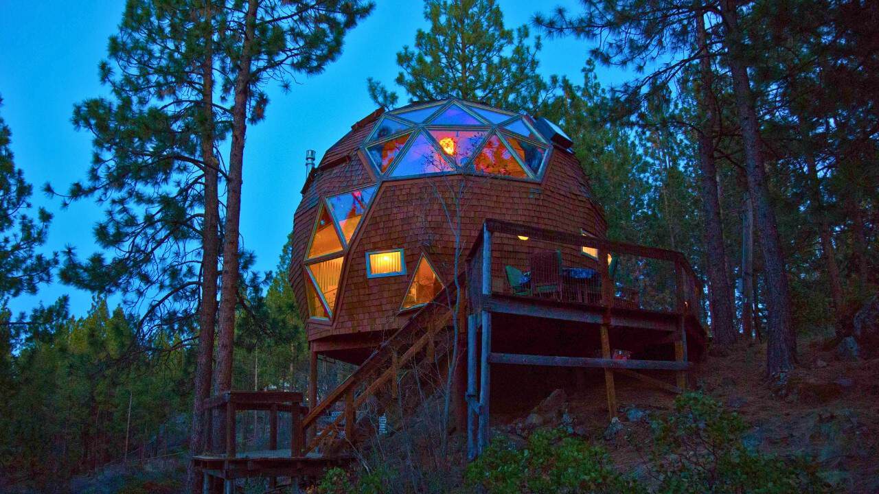 Cedar-shingle geodesic dome on a hillside timber deck at dusk, warm light in the triangular windows