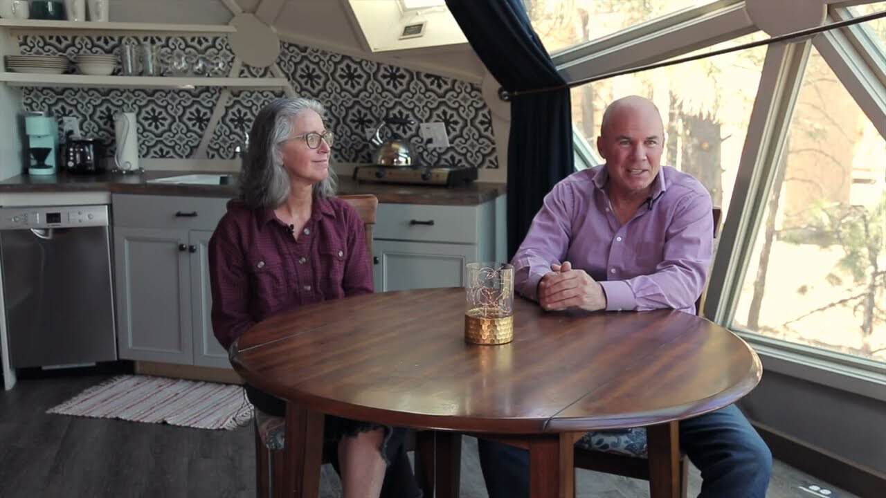 Denise and Mike at a round dining table, kitchen behind them, triangular window wall to the trees