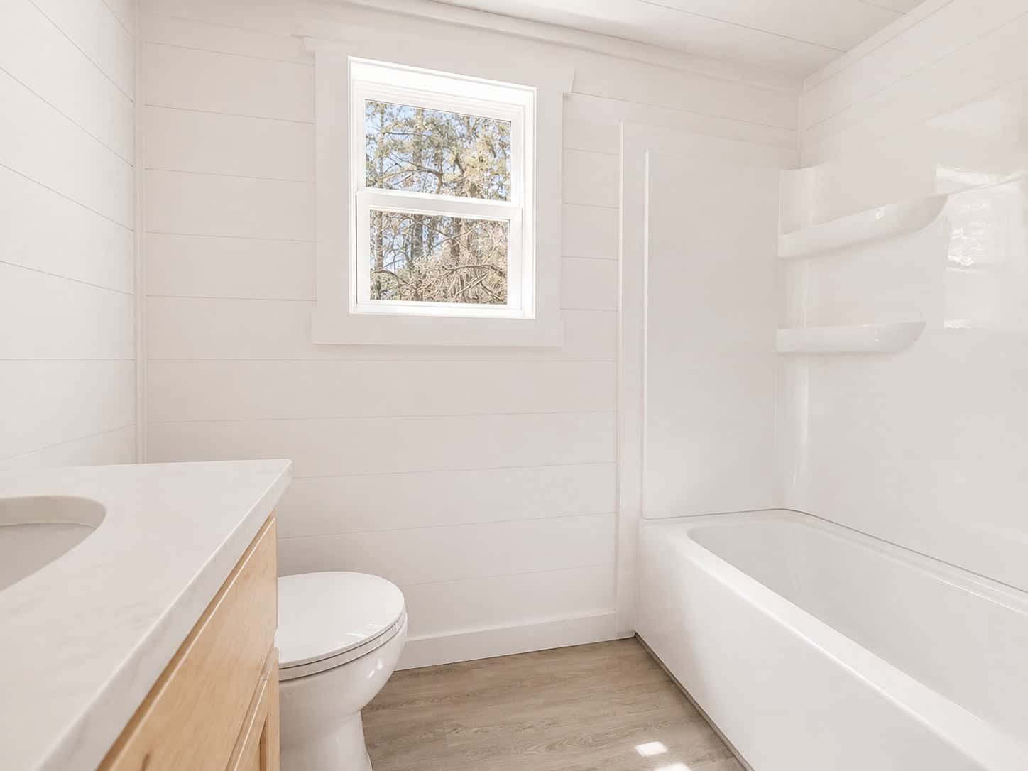 White bathroom with a tub and shower, toilet, wood vanity, and a window