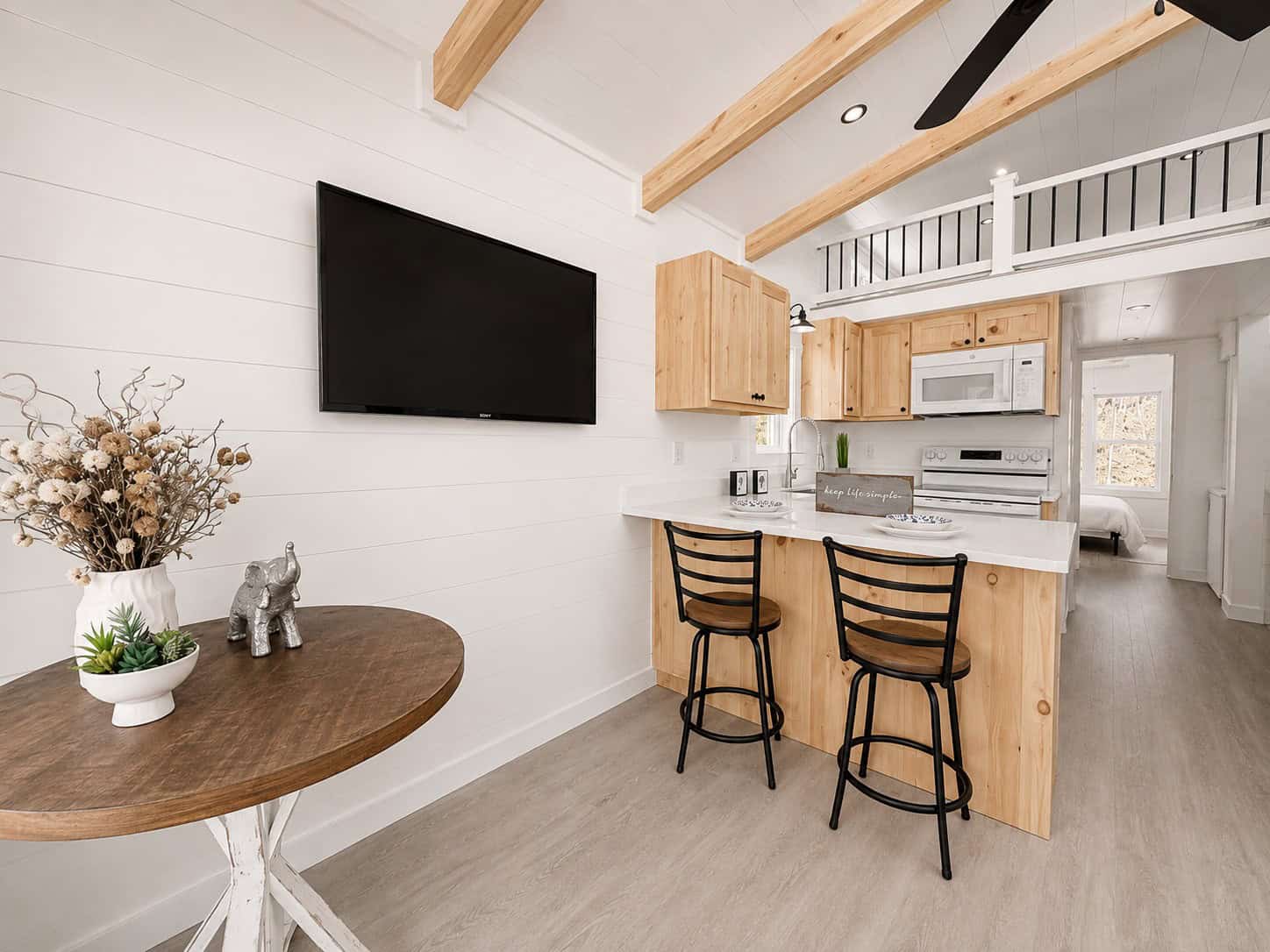 Round wood table in front of a wall-mounted TV, with the kitchen island and loft rail beyond