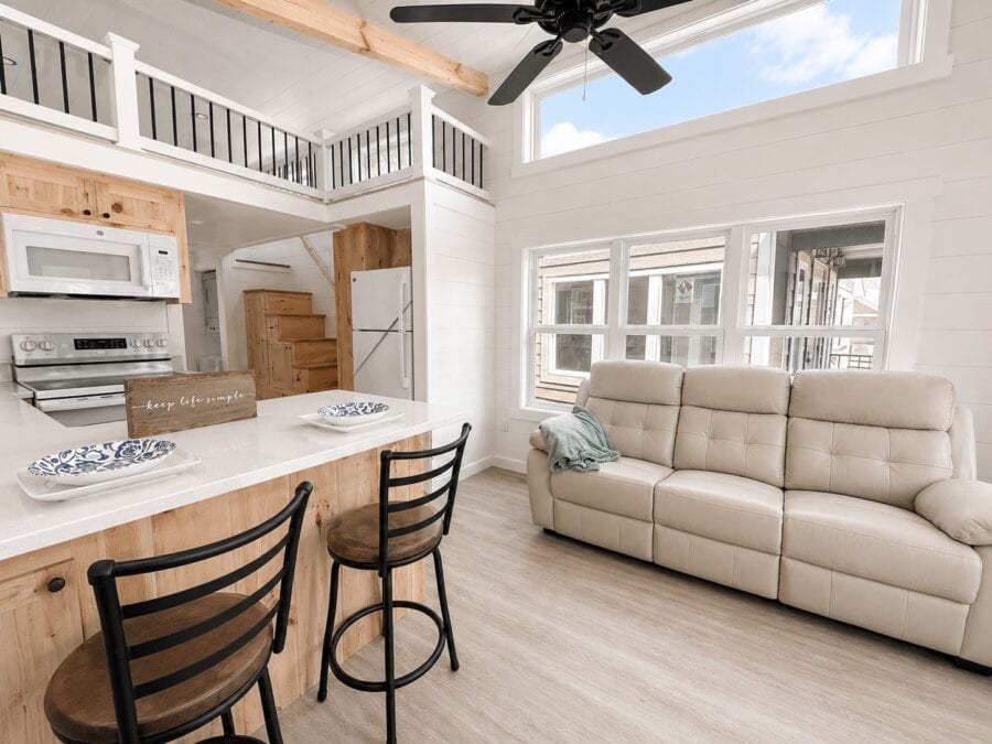 Open kitchen island with two stools, a beige sofa, high windows, and a loft rail above the kitchen