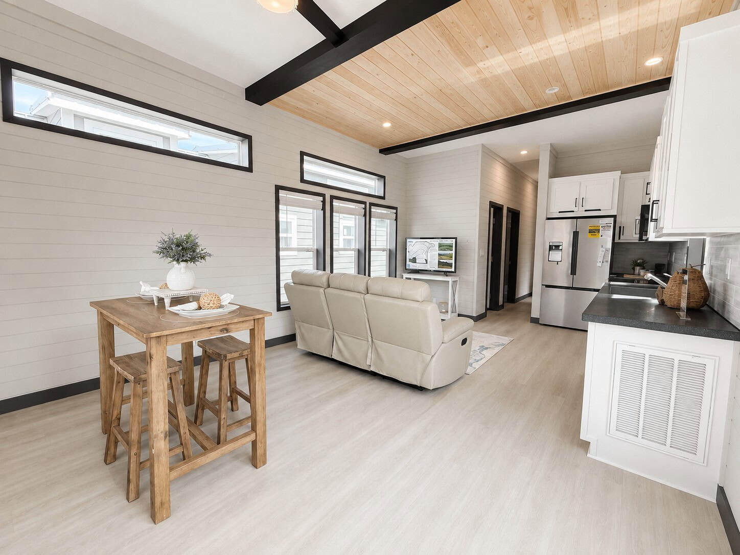 High-top wood table with stools, a beige sofa, and a kitchen run with white cabinets and a stainless refrigerator