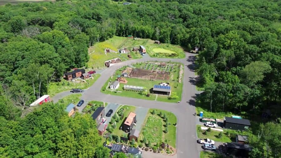Aerial overview of Tiny Timbers Agrihood in St. Croix Falls, Wisconsin: tiny homes around a loop road with a central fenced garden, greenhouse, and solar-roofed pavilion