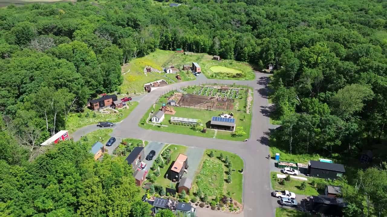 Aerial overview of Tiny Timbers Agrihood in St. Croix Falls, Wisconsin: tiny homes around a loop road with a central fenced garden, greenhouse, and solar-roofed pavilion
