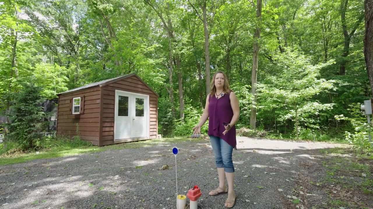 Prepared gravel lot with utility hookups and a brown 10×12 shed as founder Melissa gestures on the pad among the woods
