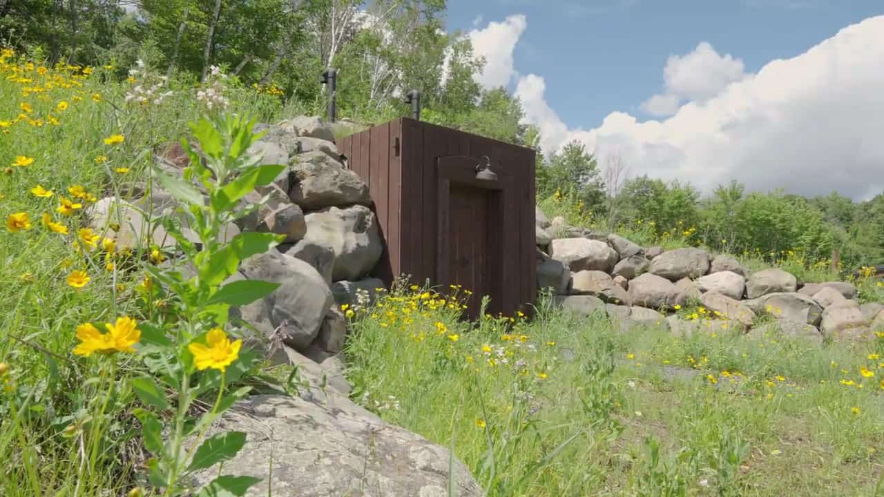 Earth-bermed root cellar with a dark vertical-plank door, gooseneck barn light, and yellow wildflowers on the mound