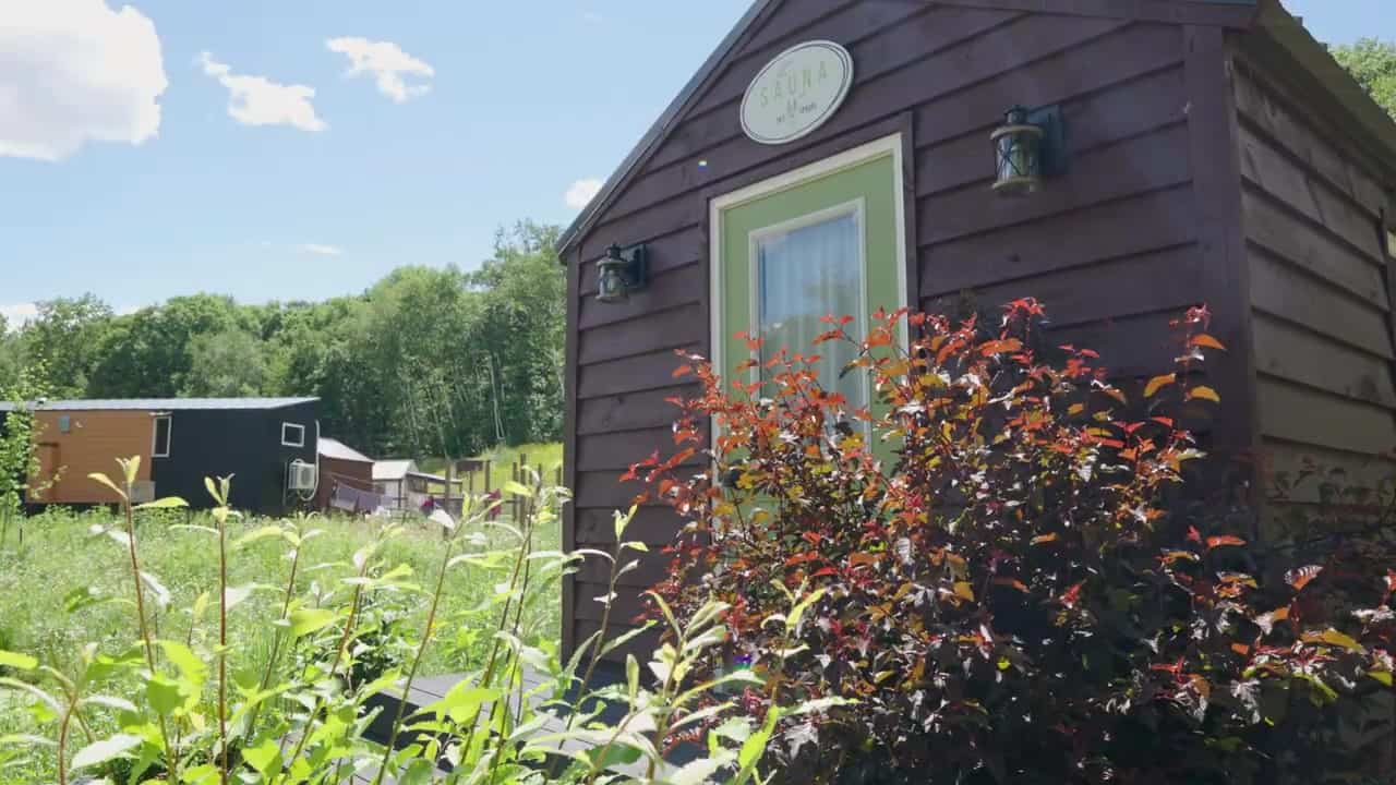 Small gabled sauna with dark brown lap siding, a light-green door, and an oval SAUNA plaque at Tiny Timbers Agrihood