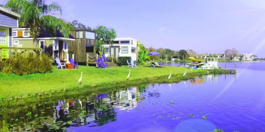 Lakefront row of tiny houses on a green lawn at Lake Fairview, Adirondack chairs on the grass, egrets at the water, yellow kayaks by a dock, at Tiny Town Orlando