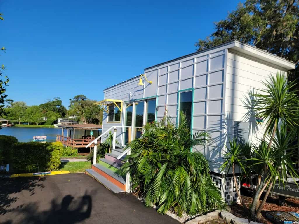 The Bermuda tiny house at Tiny Town Orlando — white siding, teal-trimmed sliders, a yellow awning, multicolor stairs, covered dock and lake behind