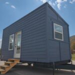 Modern tiny house on wheels with blue exterior and small wooden stairs.