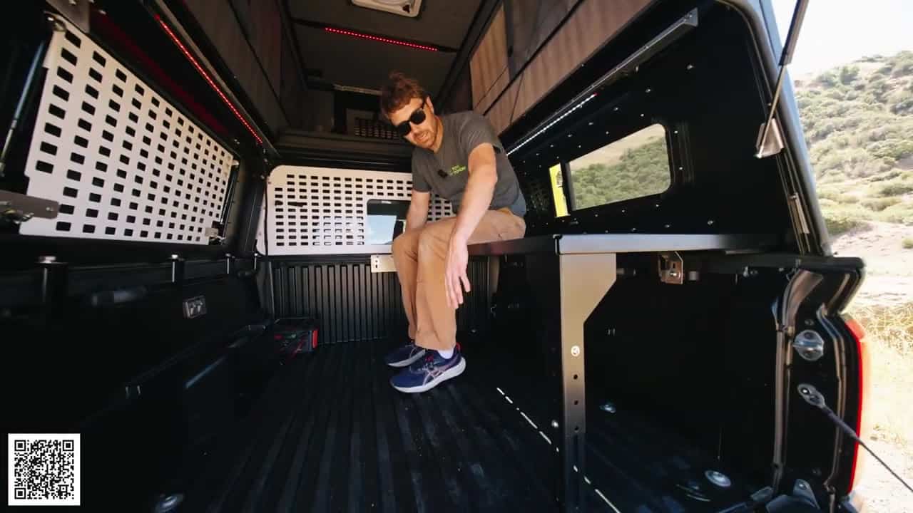 Host sitting on the optional bench and cubby inside the Badlands truck camper with white MOLLE panels across the bed