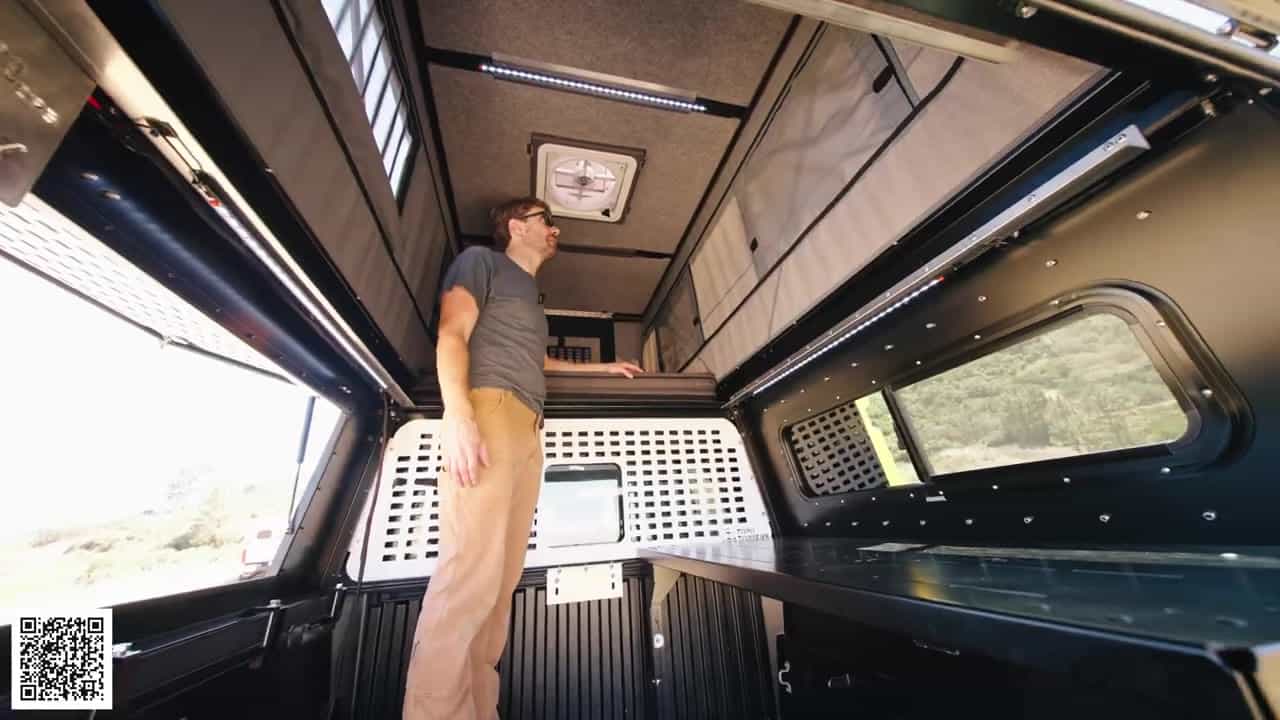 Man standing upright in the truck-bed pass-through of a popped-up Badlands camper with carpet-wrapped ceiling, Maxxair fan, and LED strips