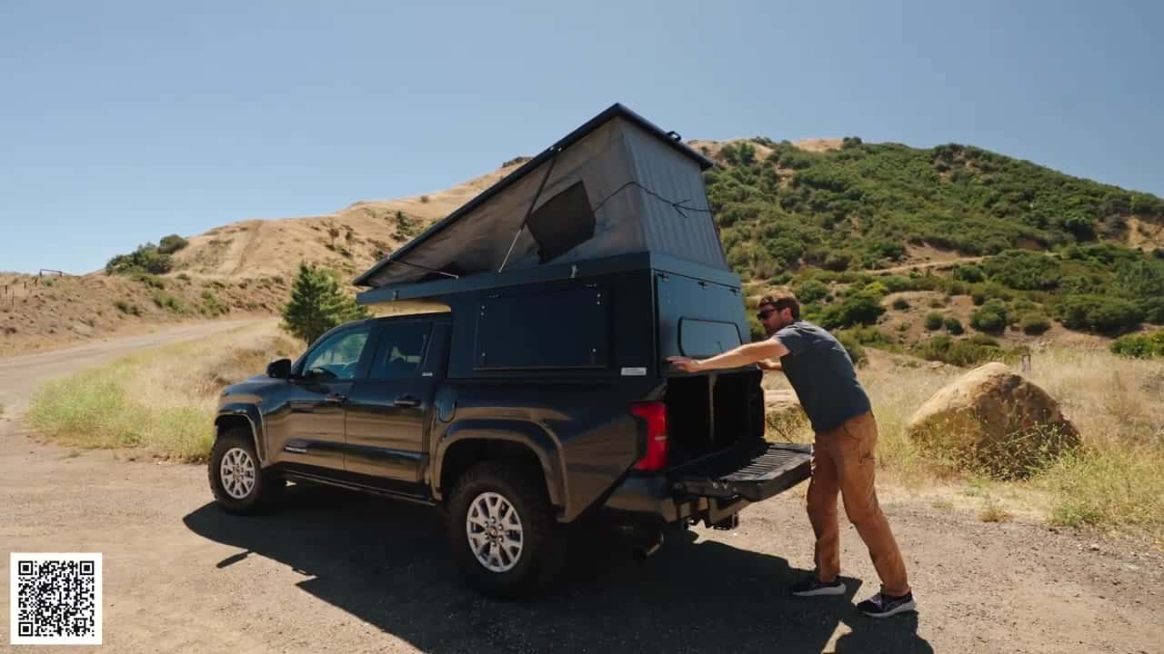 Host at the lowered tailgate of a Tacoma pushing the Badlands camper roof up into a wedge, gray tent fabric starting to open