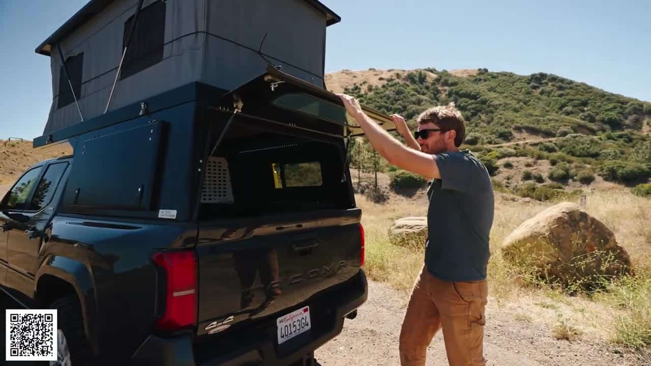 Host at the rear of a Tacoma holding the raised rear window of the Badlands camper, pop-up tent open above the black aluminum shell