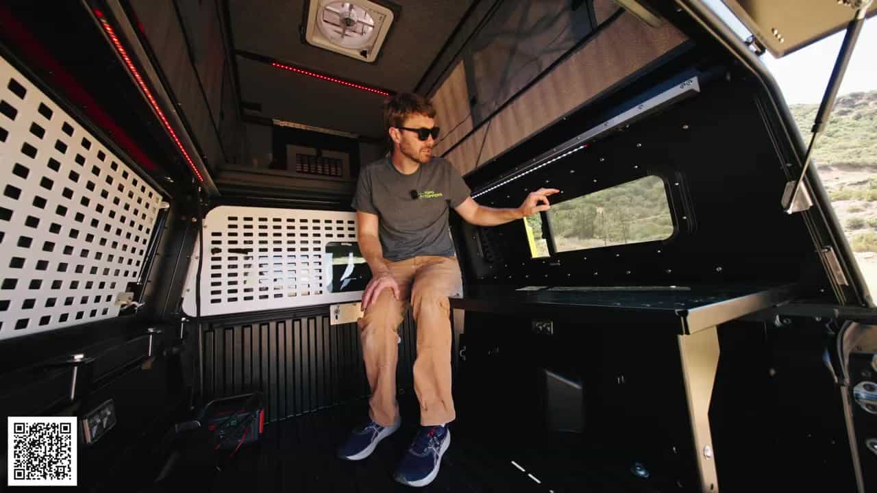 Host sitting on the interior bench of the Badlands camper pointing at a side window, white MOLLE panels on the opposite wall
