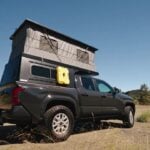 Dark gray Toyota Tacoma with a Topo Toppers Badlands pop-up aluminum canopy fully raised, gray tent walls and a yellow Rotopax on side L-track, parked on a dirt road in dry hills