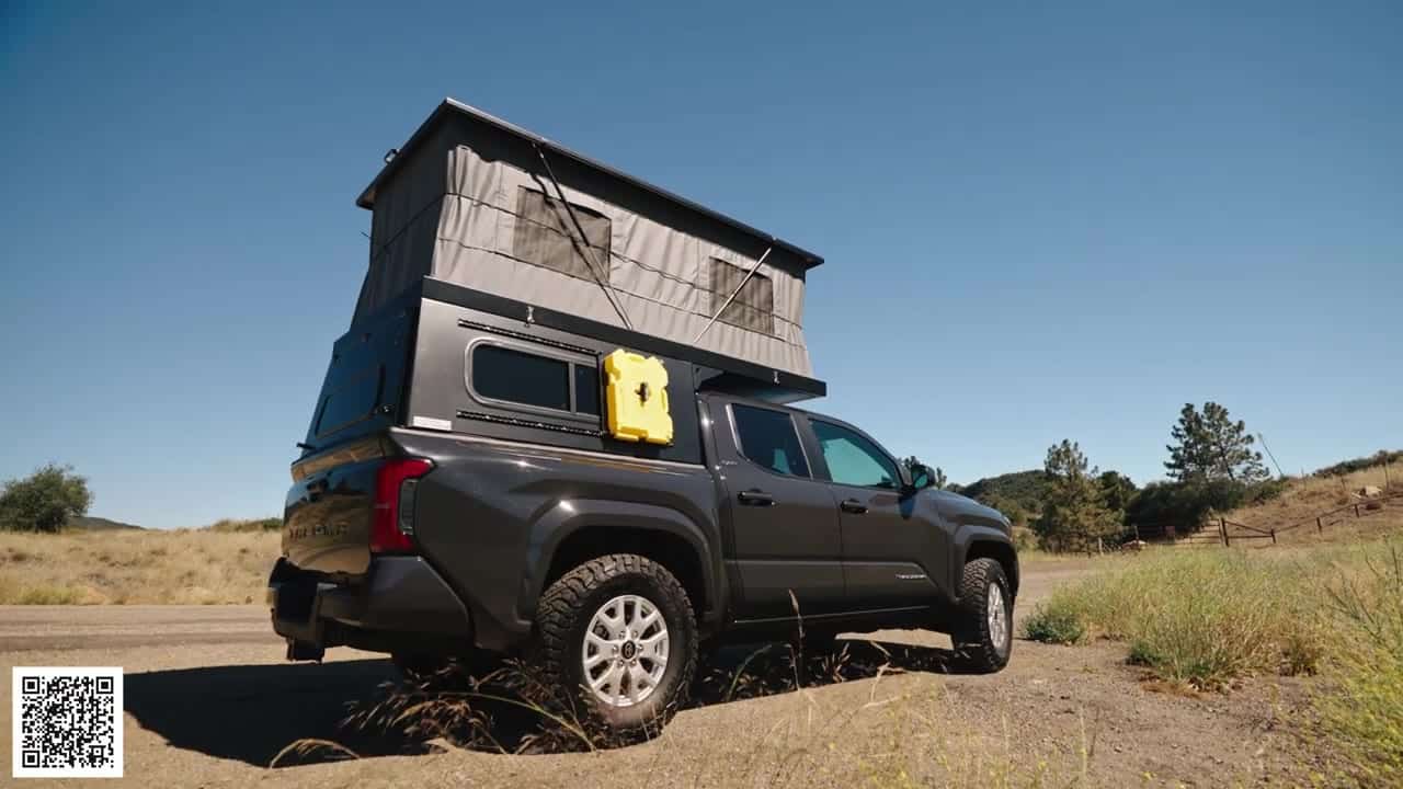 Dark gray Toyota Tacoma with a Topo Toppers Badlands pop-up aluminum canopy fully raised, gray tent walls and a yellow Rotopax on side L-track, parked on a dirt road in dry hills