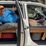 White Toyota Tundra crew cab with both driver-side doors open, a wooden platform bed inside, and Eric from ToyotorHome lying across the cab