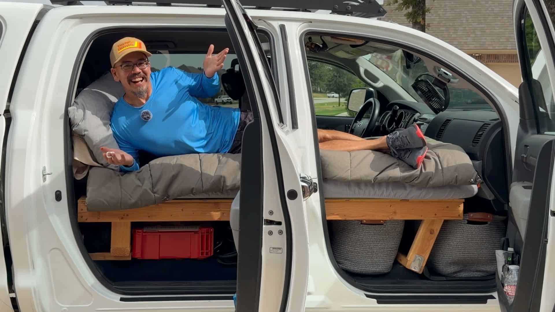 White Toyota Tundra crew cab with both driver-side doors open, a wooden platform bed inside, and Eric from ToyotorHome lying across the cab