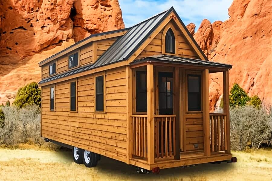 Cedar Tumbleweed Elm 26 on a dual-axle trailer: gothic porch, dark metal roof and dormer, dry grass, red rock formations under a blue sky