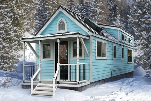 Light-blue Tumbleweed Elm 33 with white trim and a gothic-arch window over a covered porch, black metal roof, skirted, parked in snow among pines