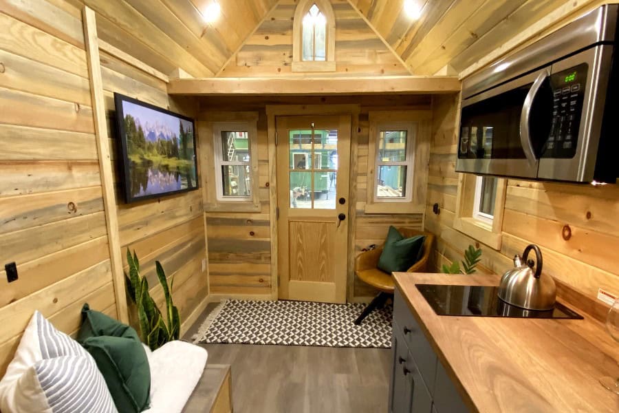 Elm great room looking toward a glass-pane door: wood walls and vaulted ceiling, two-burner cooktop and microwave, sofa, gothic-arch window above the loft edge