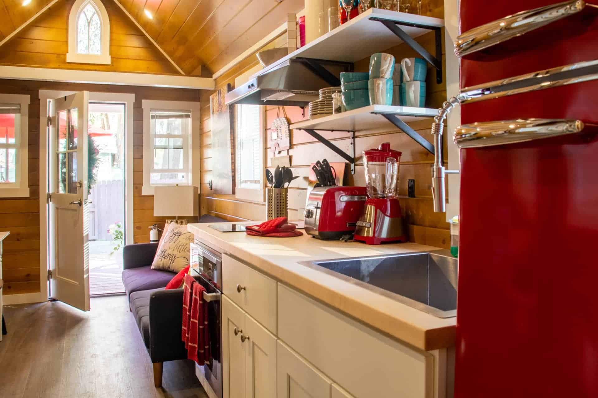 Elm interior looking toward the gothic-arch window over the porch door: red retro fridge, butcher-block kitchen, white cabinets, grey sofa, wood plank walls