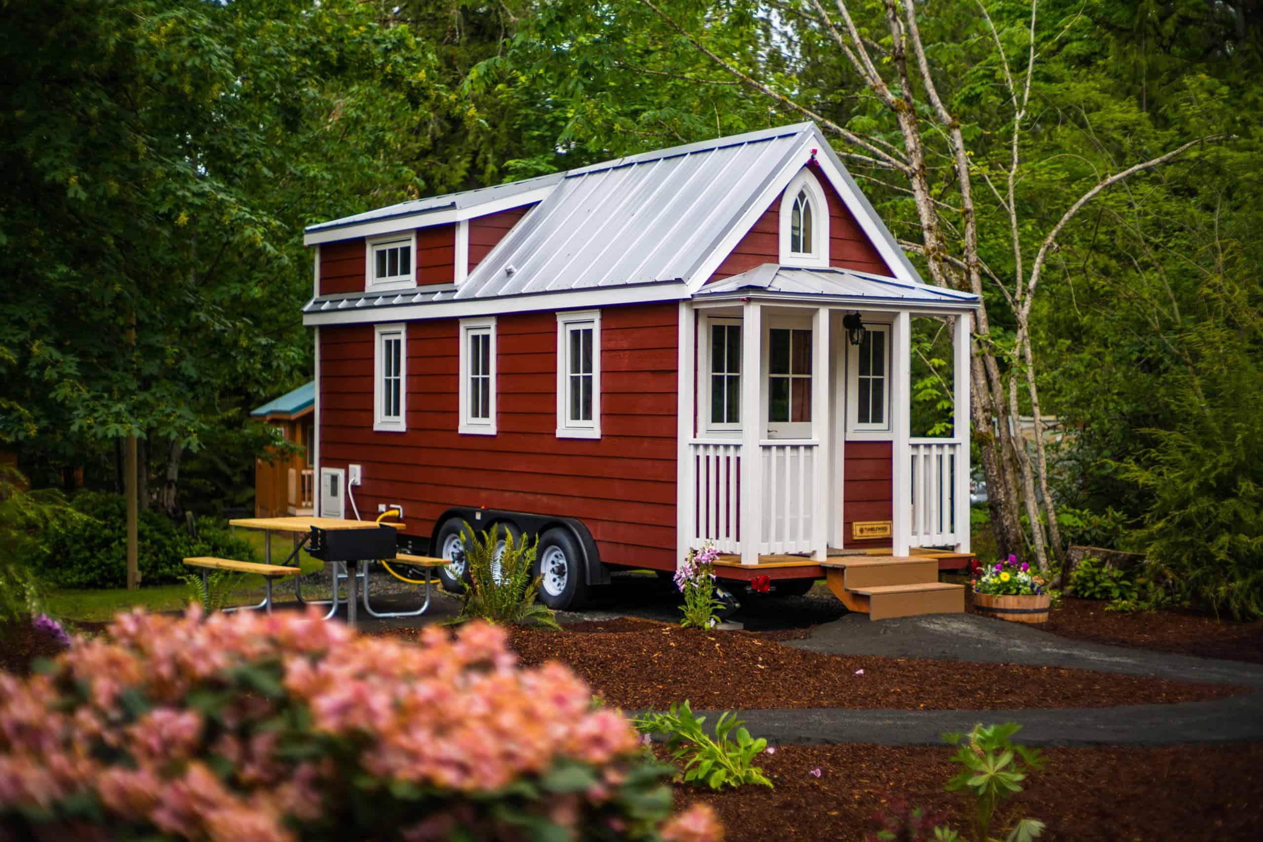 Red Tumbleweed Elm with white trim and gothic-arch window on a triple-axle trailer in the trees, covered porch, picnic table, silver metal roof